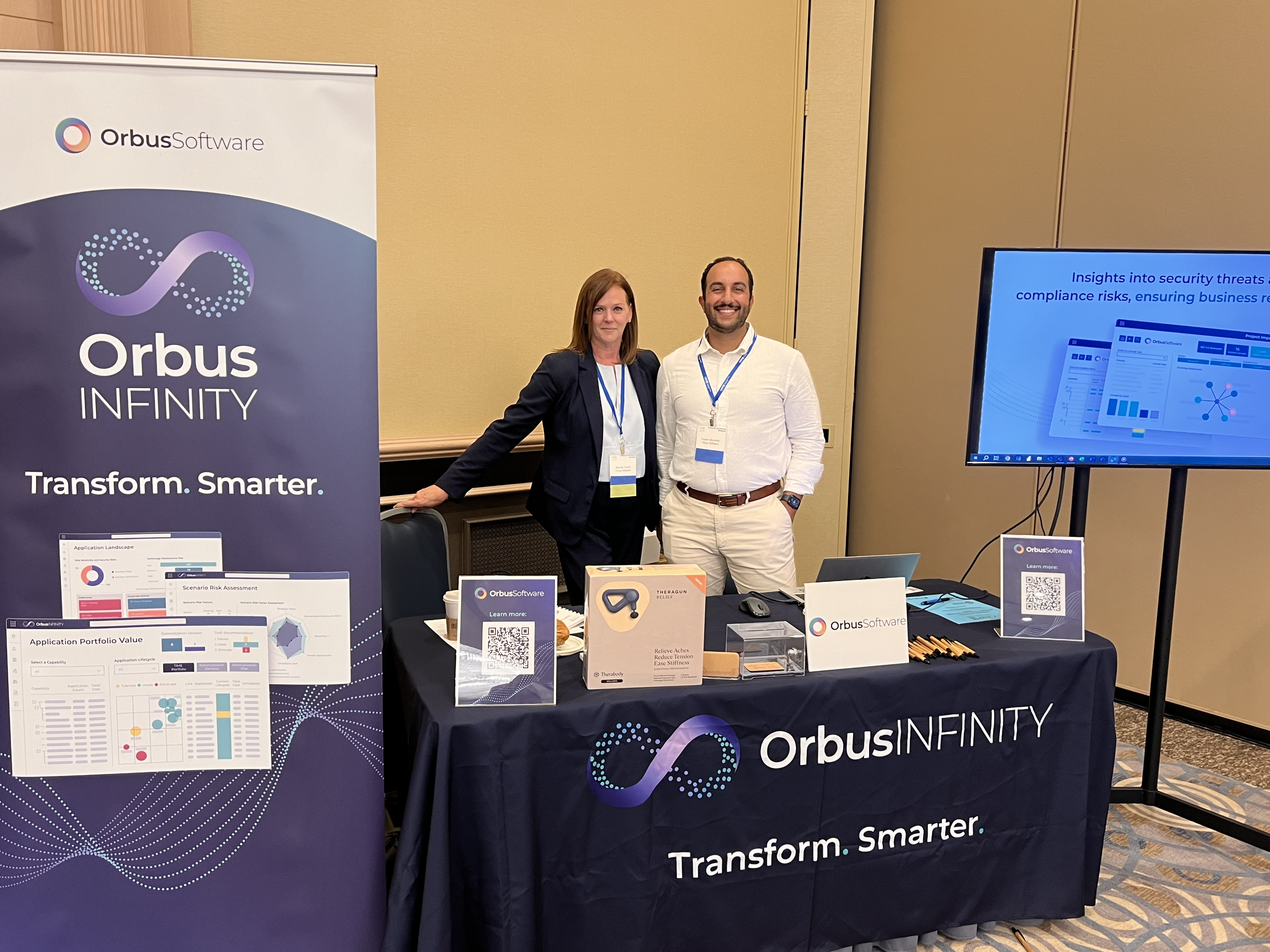 Two Orbus Software representatives, Brenda Cowie and Yazan Abutaleb, stand next to a display booth for Orbus Software at CAMP IT's 2024 Enterprise Architecture Conference.