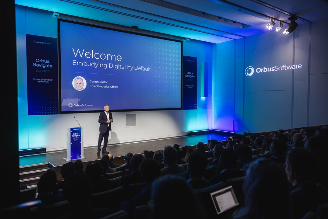 Gareth Burton, CEO of Orbus Software, presenting on stage at Orbus Navigate London event. The screen behind him displays 'Welcome' and 'Embodying Digital by Default,' along with his name and title.