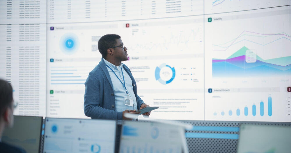 A man standing in front of a large digital screen displaying various charts, graphs, and data visualizations.