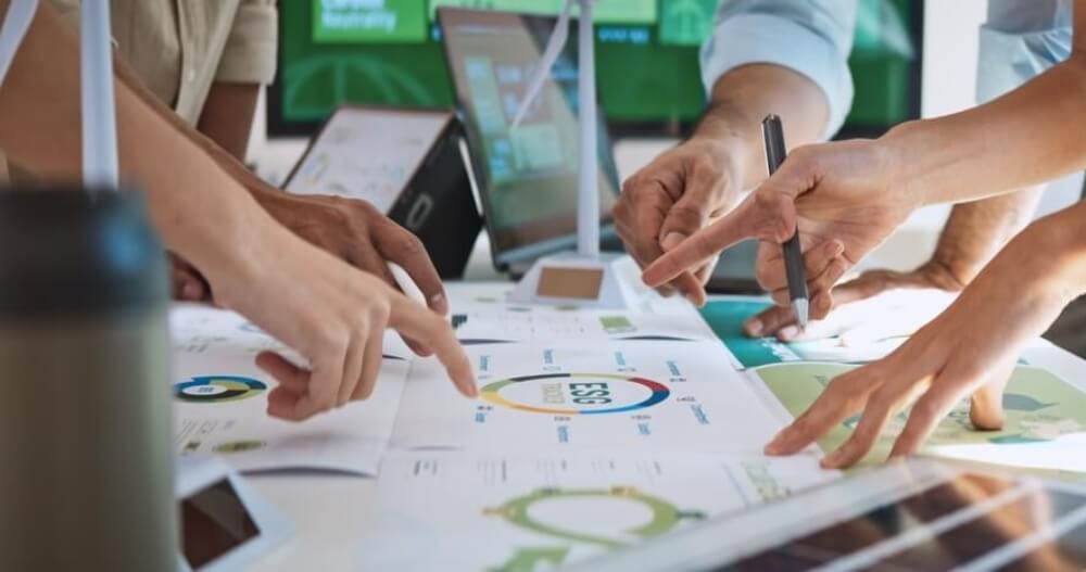 Several hands point and gesture toward documents on a table displaying graphs, charts, and diagrams related to ESG or sustainability.